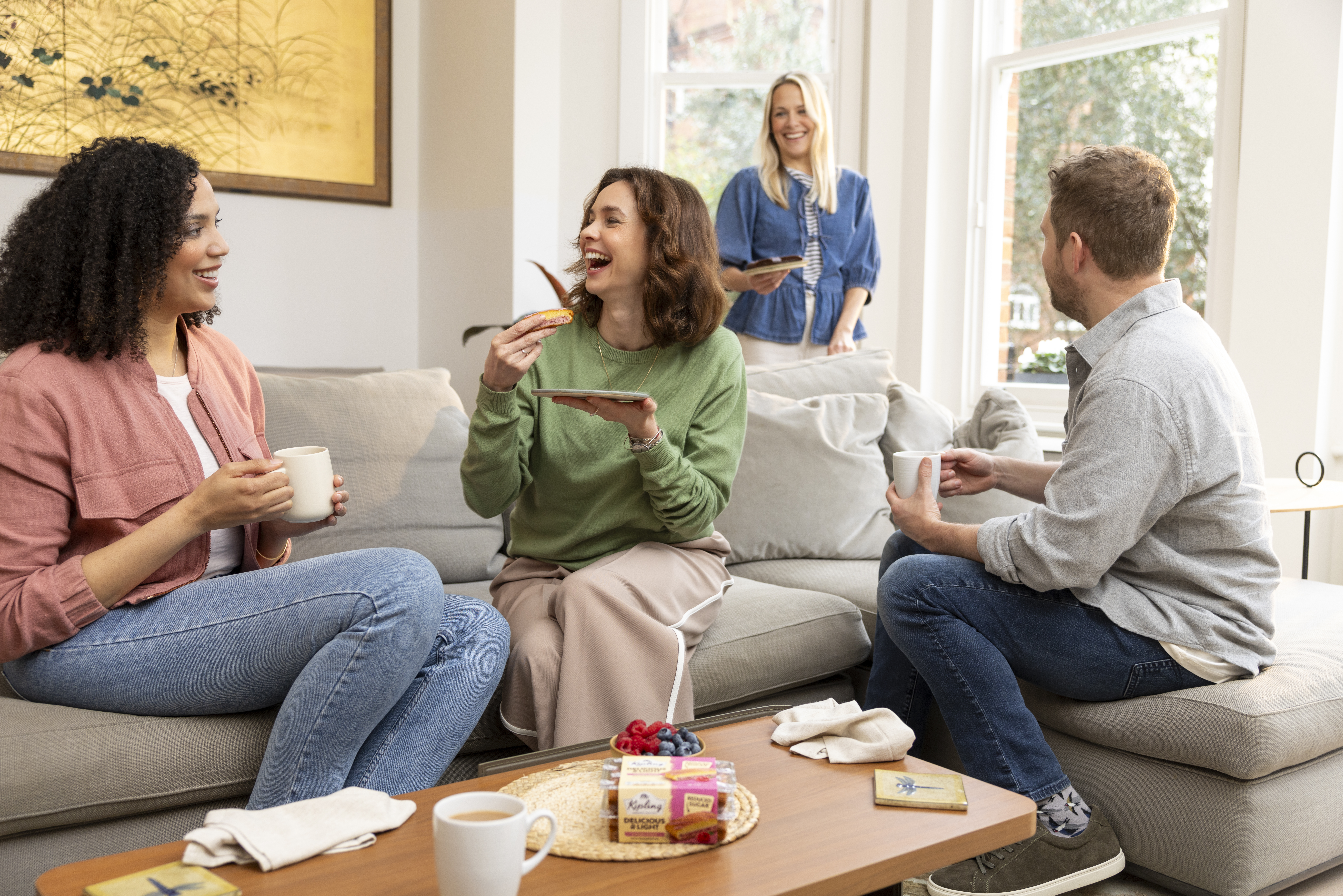 Four friends gathered around a sofa with hot drinks and cake slices